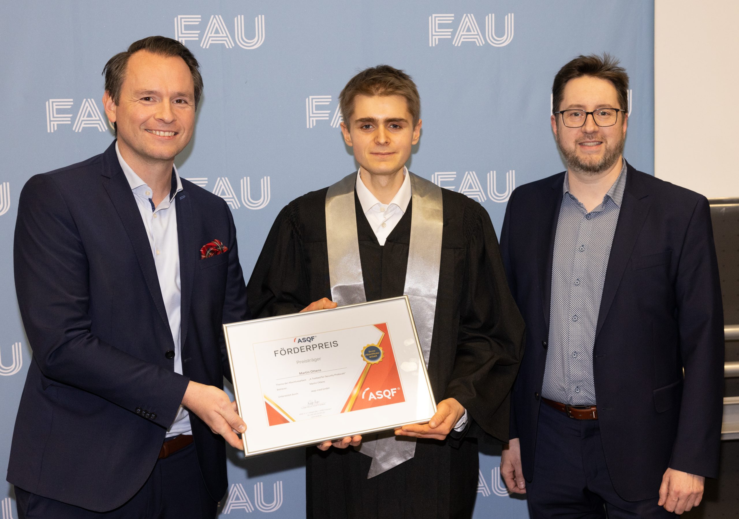 Three men stand in front of an FAU backdrop. The man in the middle, wearing a graduation gown, is holding a framed certificate. The two men next to him are dressed in business attire and smiling at the camera.