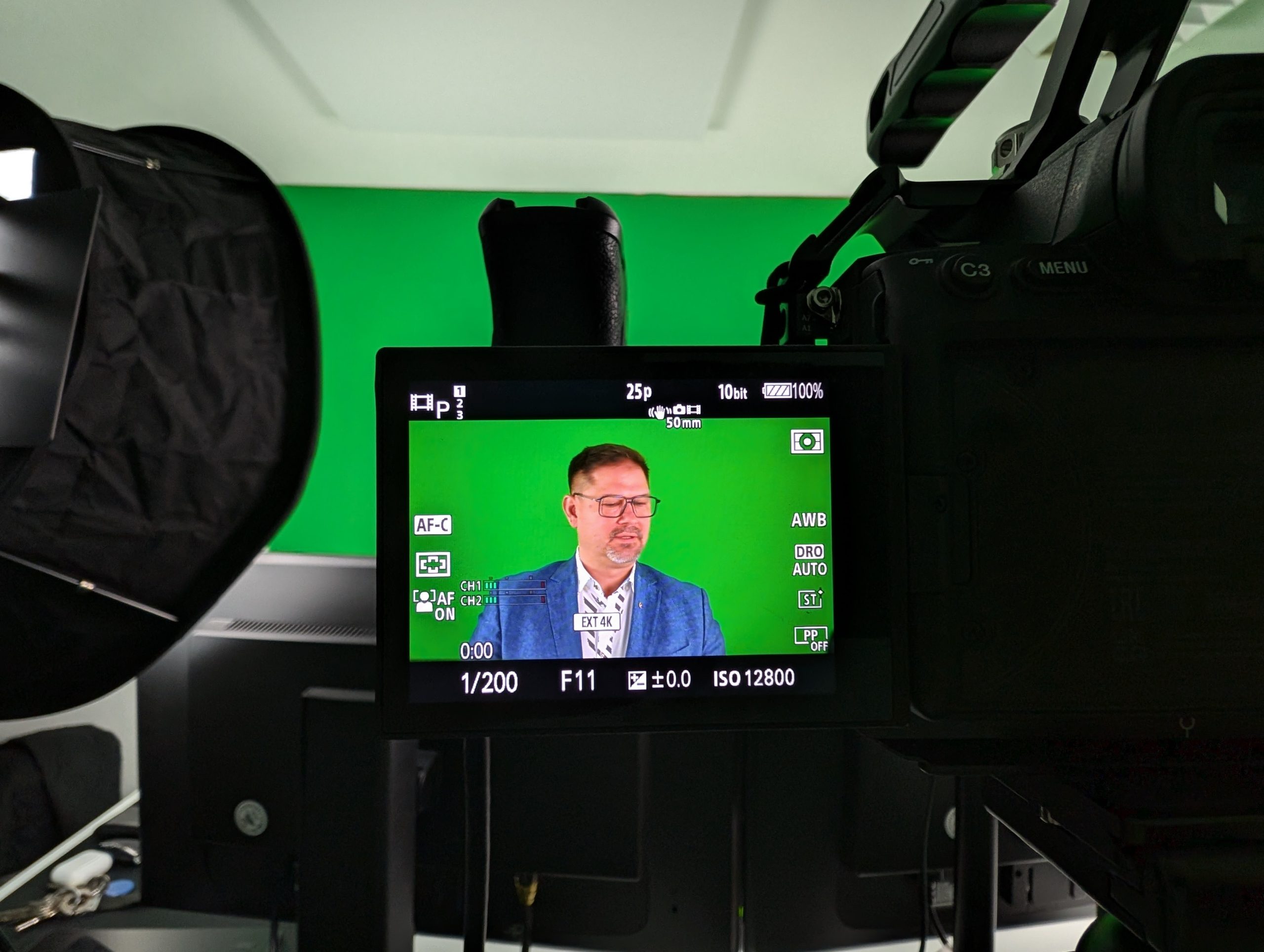 A man in a blue suit can be seen on a camera screen speaking in front of a green screen in a studio. Professional lighting and recording equipment can be seen around him.