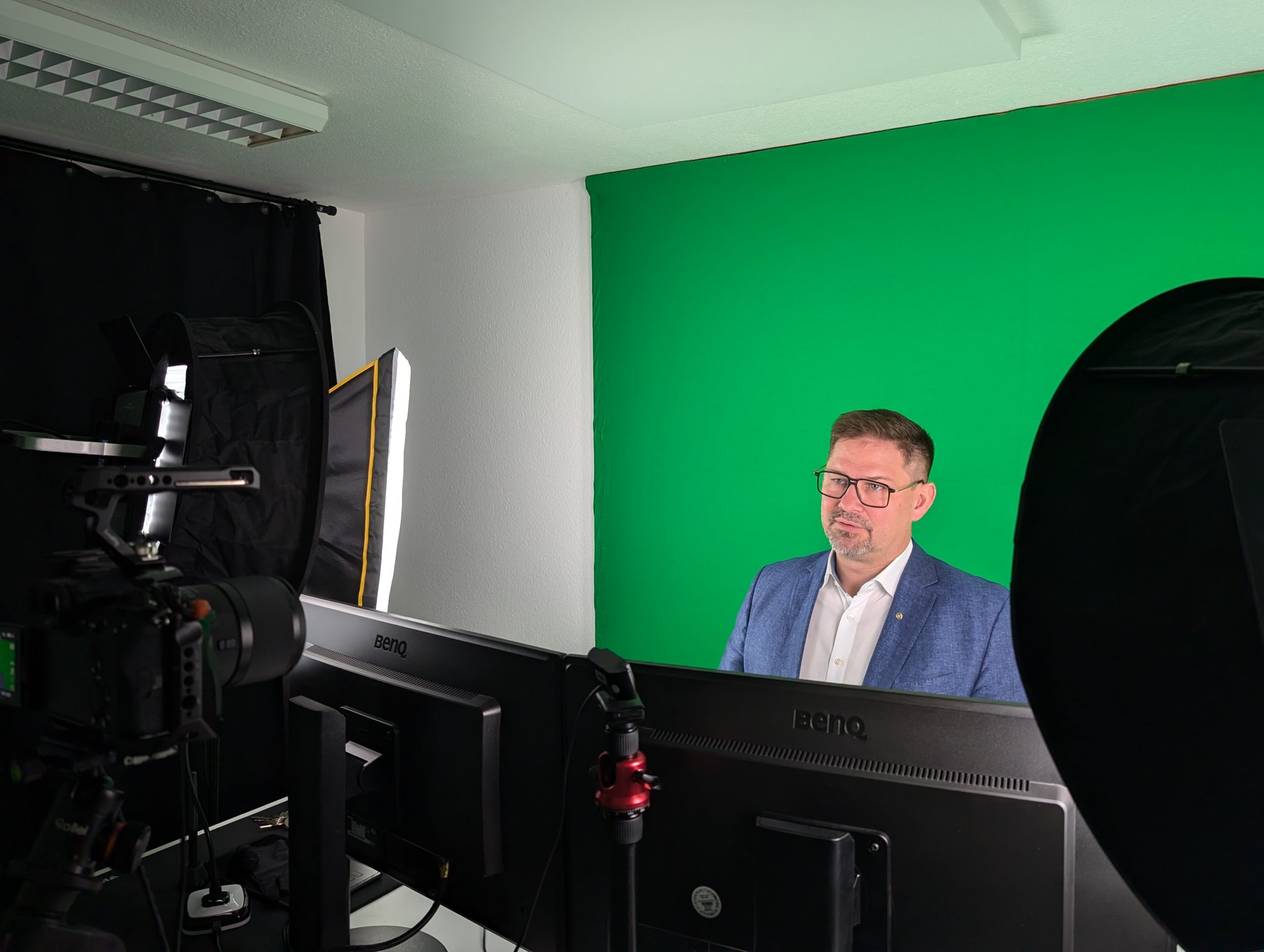 A man in a blue blazer sits in front of a green screen, surrounded by studio lights, monitors and camera equipment, preparing for a video recording or livestream.