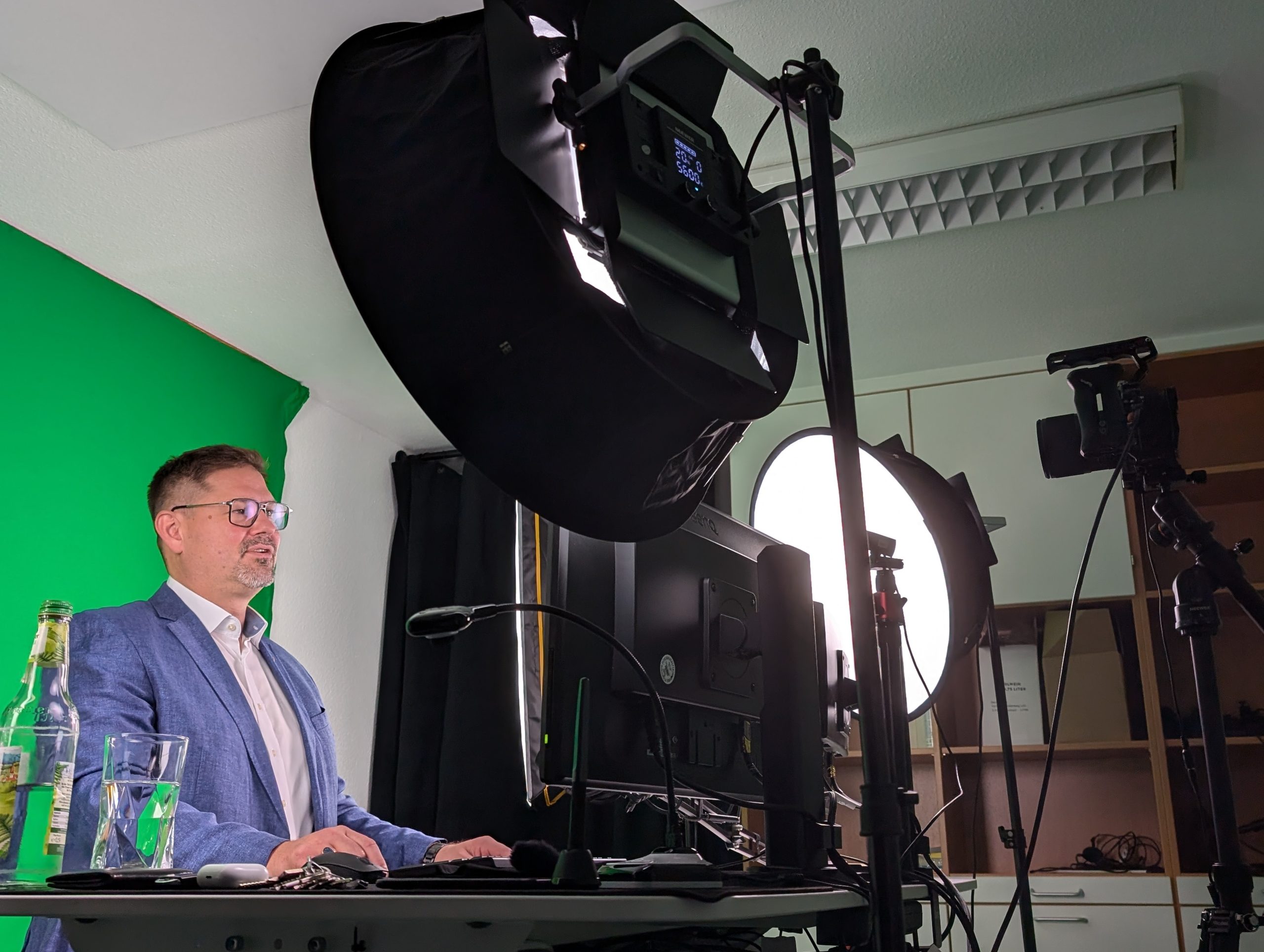 A man in a blue blazer is sitting at a desk with a computer in front of a green screen and large studio lights, preparing for a video recording or livestream. A camera and a water bottle can also be seen on the desk.