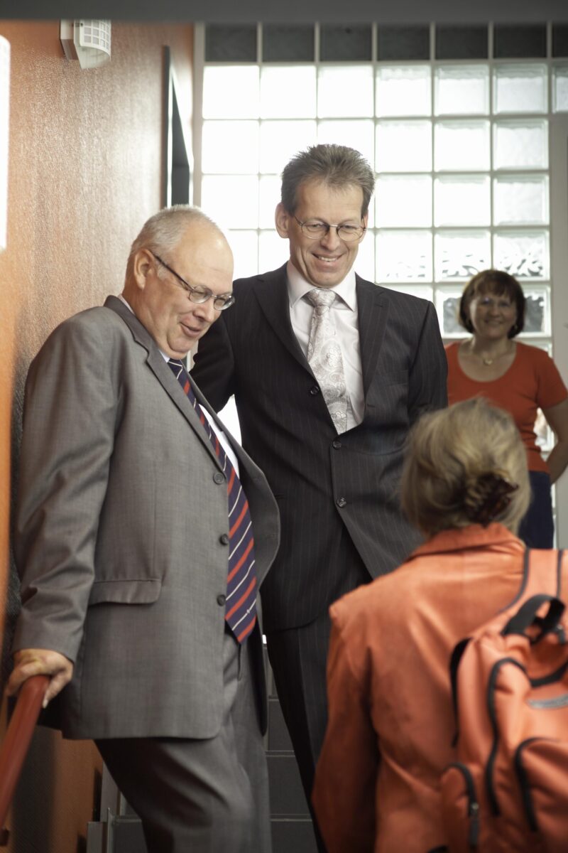 Two men in suits stand on a staircase, smiling and talking, while a woman in a red jacket with a rucksack stands opposite them. Another woman in a red top is standing in the background and is also smiling.