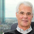 An older man with white hair and glasses sits in an office chair and smiles, while a cityscape and a tall building can be seen through the window behind him.