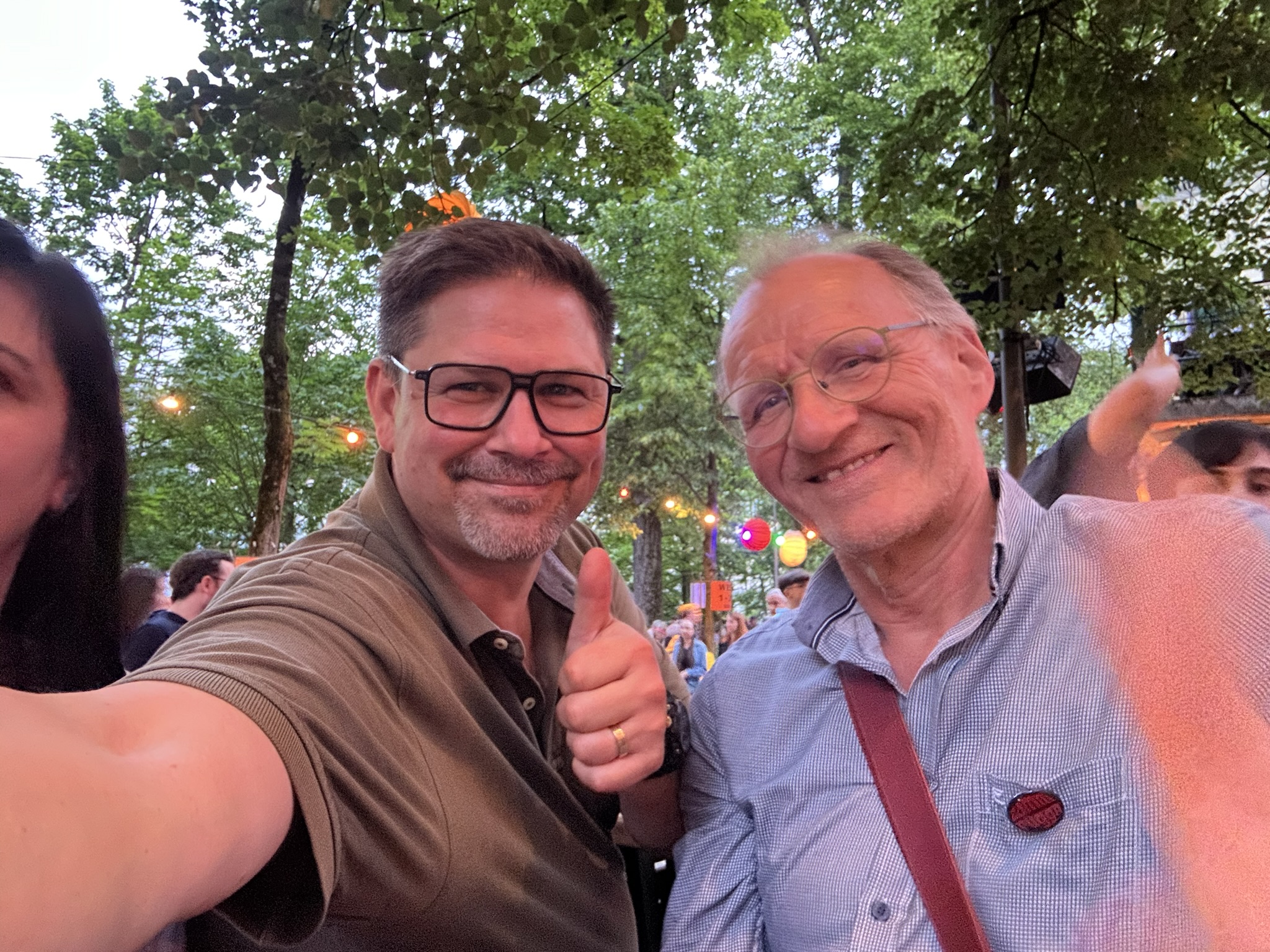 Two smiling men pose for a selfie at an outdoor event with trees and string lights in the background. One man gives a thumbs up, and people can be seen in the festive, colorful setting behind them.