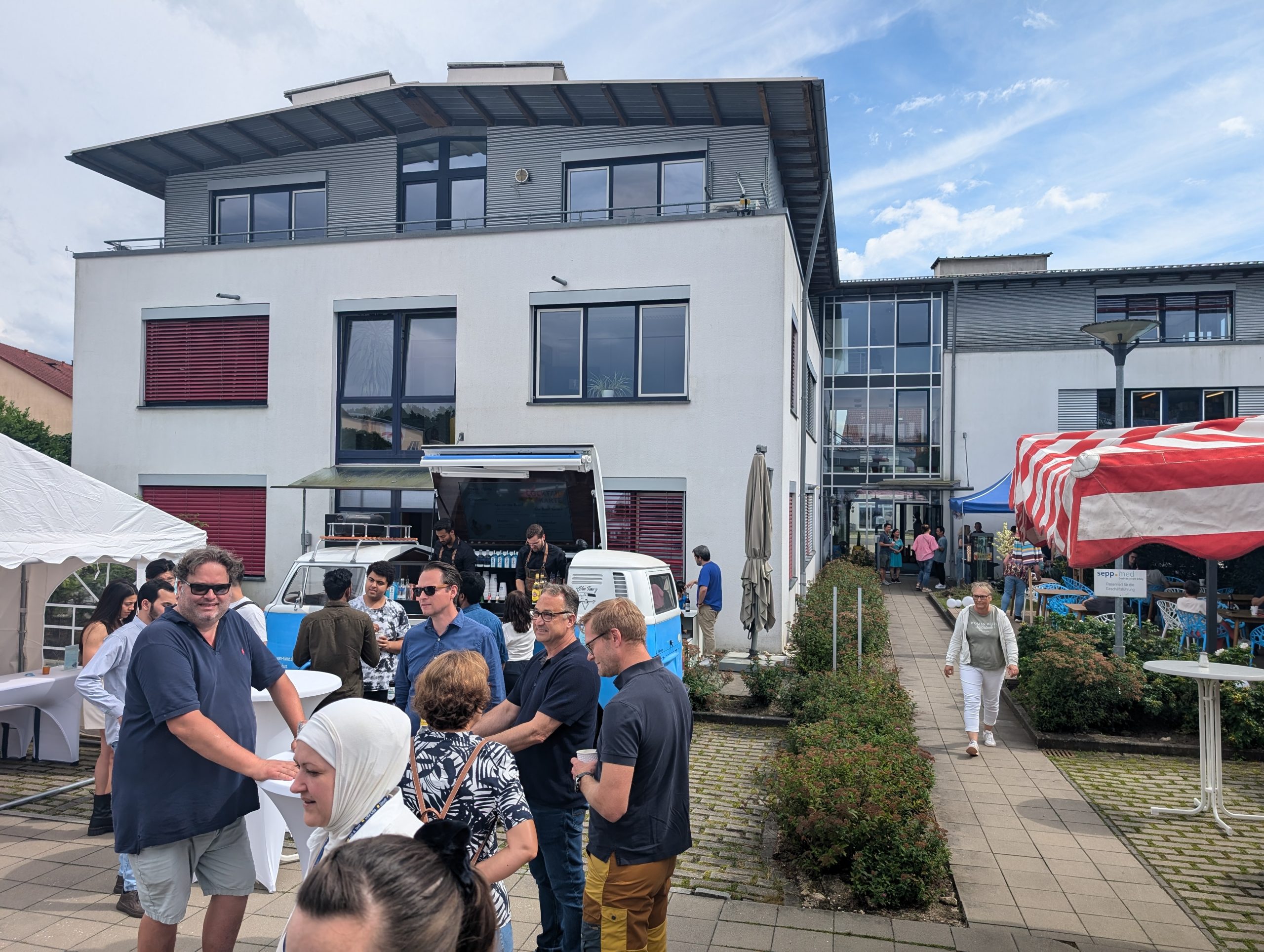 A group of people gather and chat outside in front of a modern white building with market tents and a food truck. A few people are standing in a row and the sky is partly cloudy.