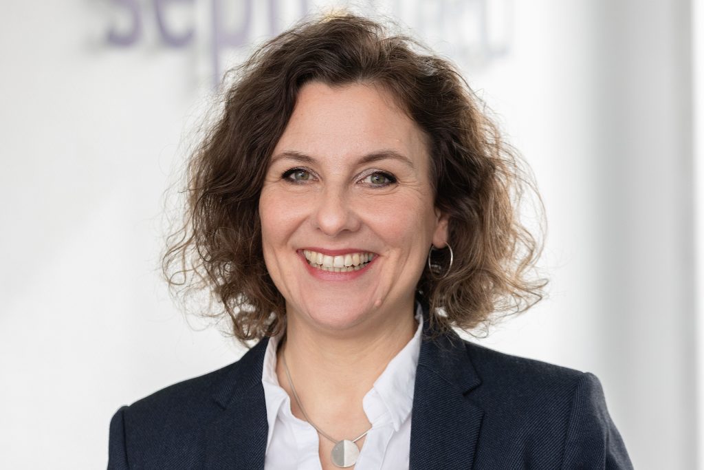 A woman with curly brown hair, wearing a dark blazer over a white blouse, smiles at the camera. She is standing in a bright interior with a blurred background.