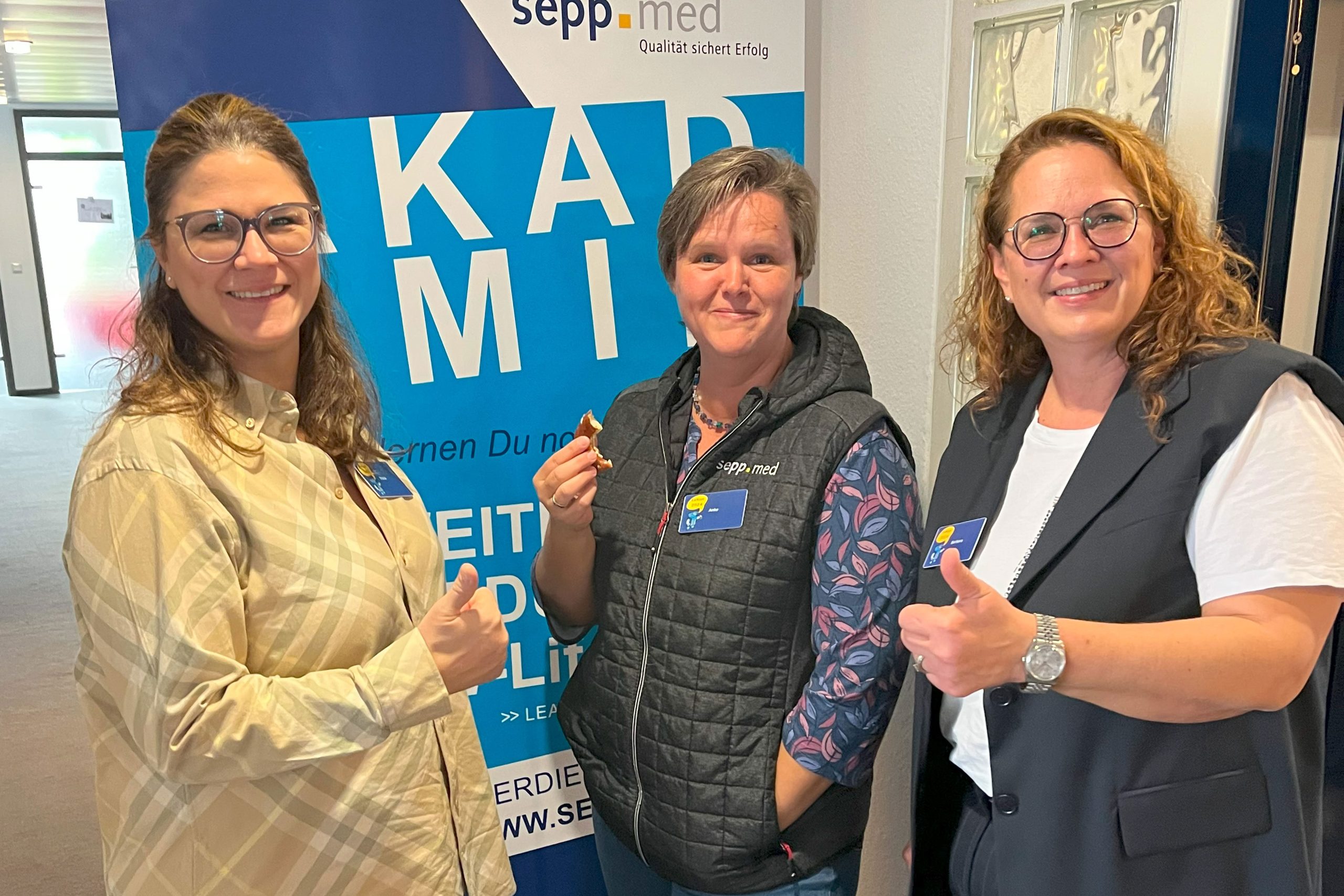 Three women, smiling and giving a thumbs-up, stand in front of a blue and white sepp.med banner. They are wearing name badges and casual business attire in a well-lit interior.