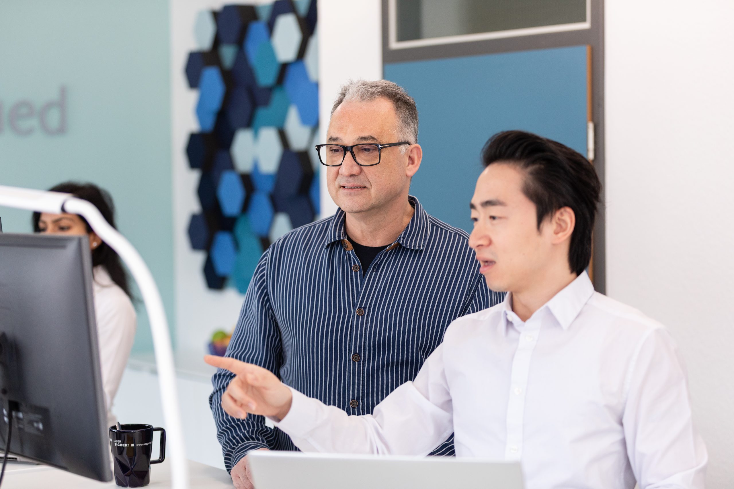 Two men are working together on a computer in an office. One, seated, points at the screen while the other, standing next to it, watches attentively. Geometric wall art and a door can be seen in the background.