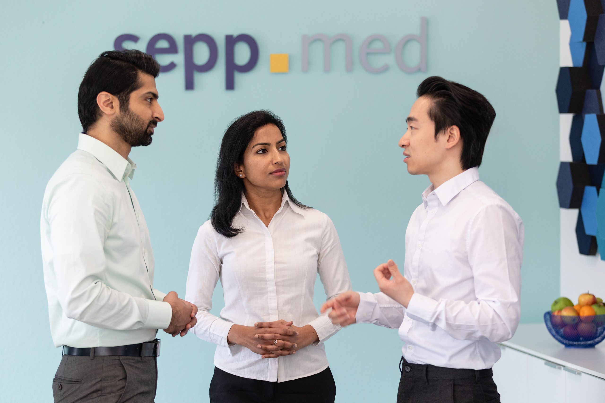 Three professionals in business attire stand and have a discussion in an office with a sepp.med logo on the wall and a bowl of fruit on a counter in the background.