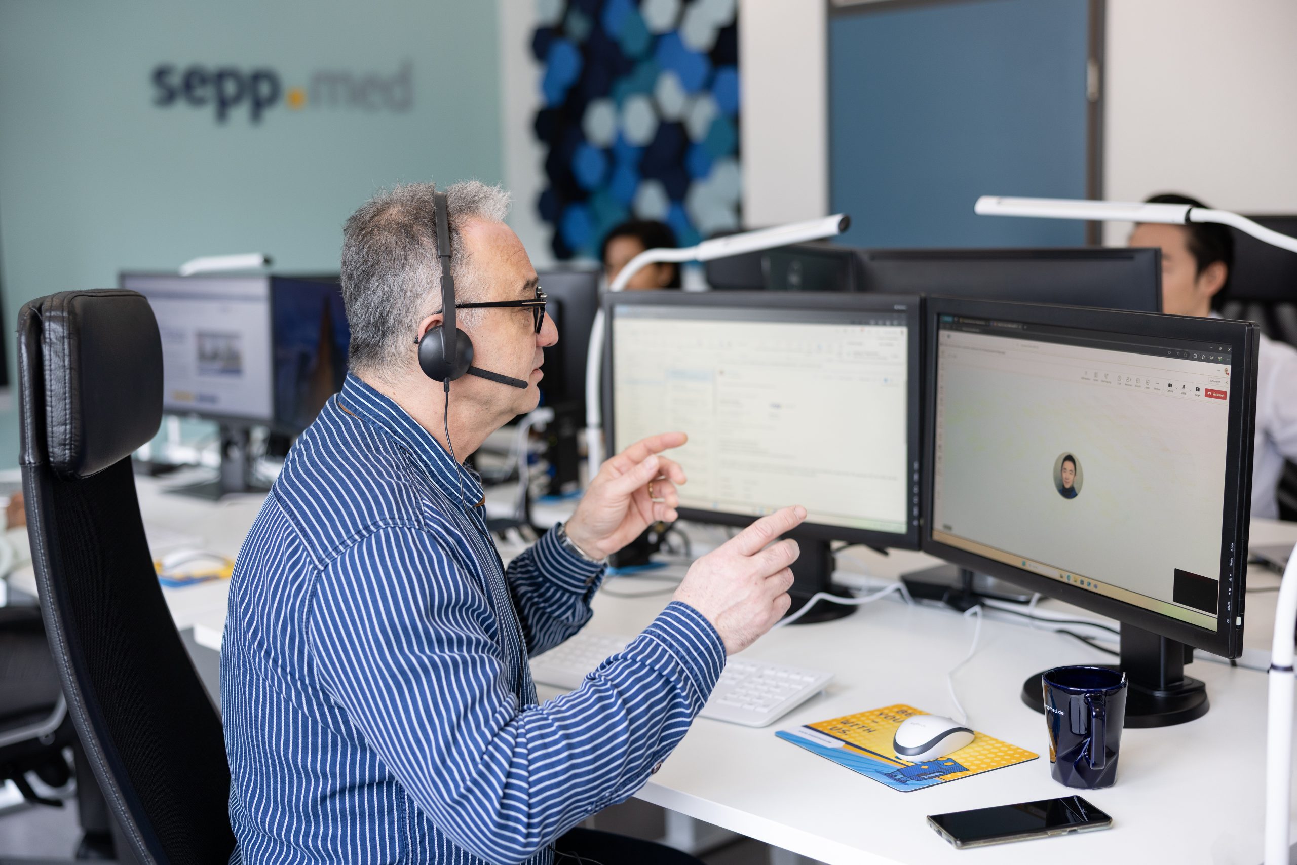 A man wearing a headset sits at a desk with two monitors, engaged in a video call or online meeting in a modern office setting. A coffee mug, keyboard, and mouse are on the desk. Other people are in the background.