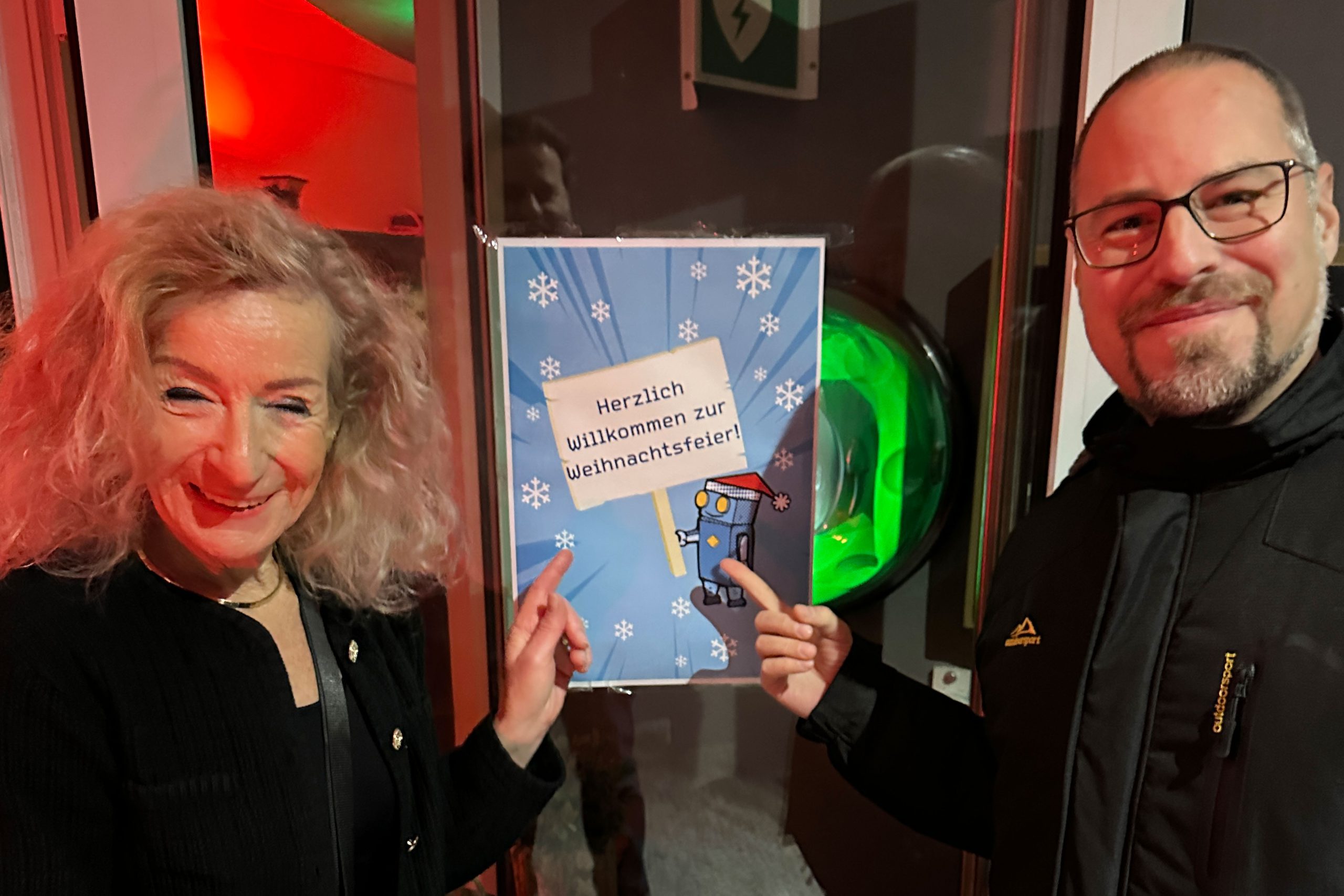 Two smiling adults stand next to a festive sign saying "Welcome to the Christmas party!" with snowflakes and a cartoon character in a warmly lit room.